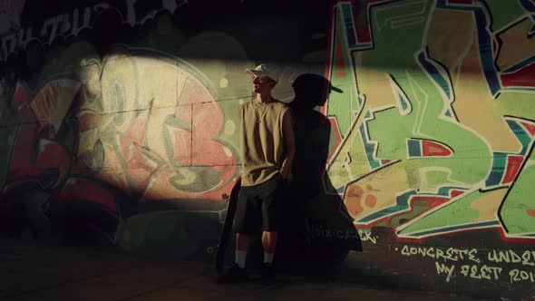 Young Hipster Resting with Skate Board at at Sun Rays Near Graffity Wall alt