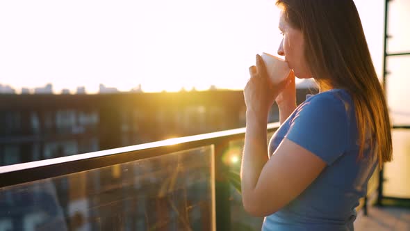 Woman with a Cup of Coffee Standing on the Balcony and Admire the Sunset alt