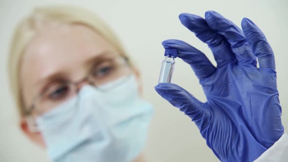 A Female Doctor in a Medical Mask and Rubber Gloves Holds a Small Ampoule with a Blue Drug in Her