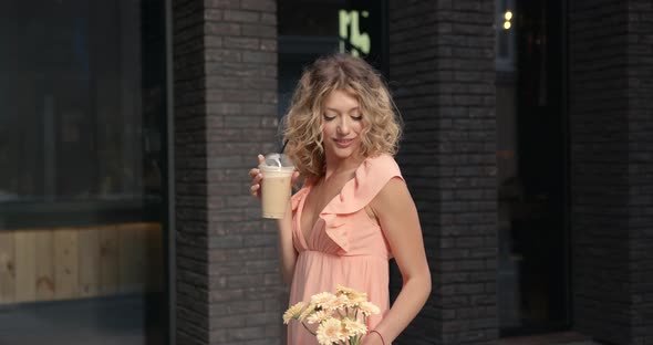 Smiling Woman Standing Outside Drinking a Coffee and Carrying Flowers alt