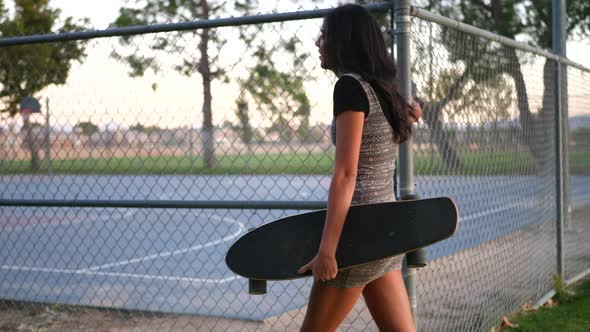 A pretty young hispanic woman smiling and walking through an urban city park with her skateboard alo alt