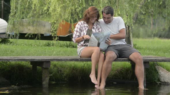 Young Smiling Couple in Nature Look at Map Seated on Lake Jetty in Summer Day Outdoor alt