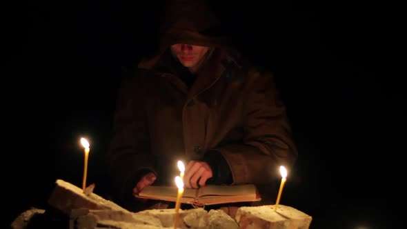 a Man in a Cloak with a Hood is Reading a Hot Book with Candles on the Floor alt
