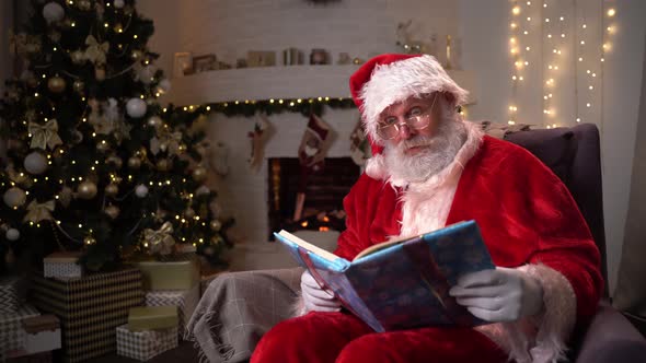 Funny Santa Clause Sitting Near Fireplace and Christmas Tree Reading a Magical Book with Shining
