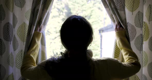 Rear view of woman at home opening curtains and looking out of the window