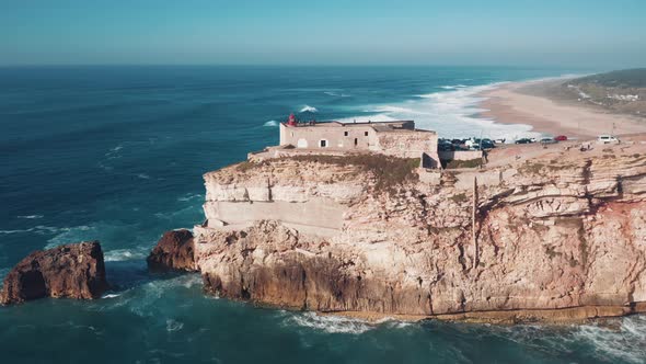 Aerial view of Nazare seascape, old Fortress and lighthouse on cliff; blue stormy water of Atlantic