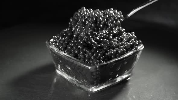 A Man Scoops Up a Delicious Black Caviar From a Transparent Bowl with a Spoon alt