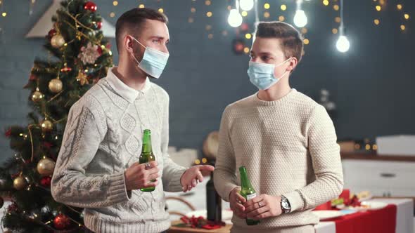 Two Beautiful Young Guys in Medical Masks Celebrate the New Year. A Table and a Christmas Tree Are alt