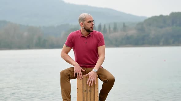 Pensive musician playing cajon in nature against lake and mountains alt