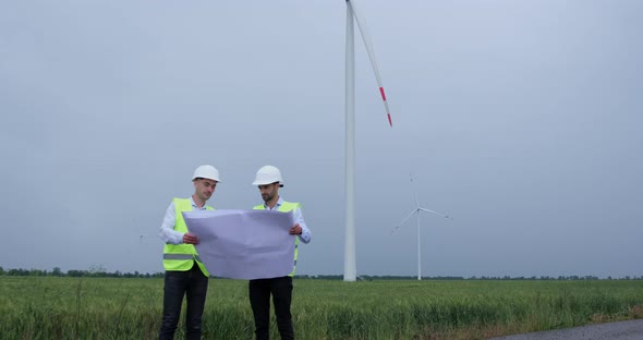 Engineers look at project sheet in hands and point to wind turbine generating electricity.