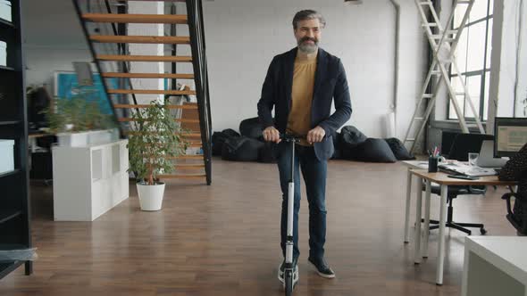Mature Businessman Riding Scooter in Workplace Smiling While People Working at Tables alt