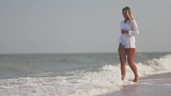 A Slender Girl in a Gentle Blue Swimsuit and Shirt Walks Along the Sandy Beach Near the Blue Sea alt