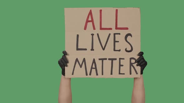 Female Hands in Black Gloves Pick Up a Poster From a Cardboard Box That Reads ALL LIVES MATTER alt