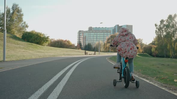 Little Girl Rides a Tricycle Bike in the City Park at Sunset in Sunny Autumn alt