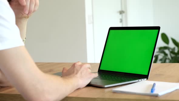 Close-up of a Man Uses Laptop with Green Mock-up Screen While Sitting at the Desk alt