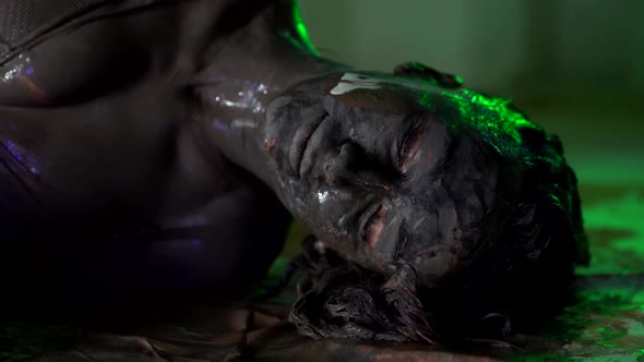 Creative Shooting of Woman with Paints on Face and Body Dirty Model Is Lying on Floor of Studio alt