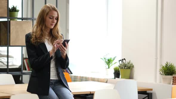 Browsing Online on Smartphone by Businesswoman Sitting on Desk alt
