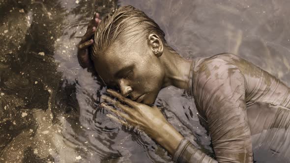 Unusual Fashion Shot with Tender Woman Covered Golden Paint in Water Moving Slowly alt
