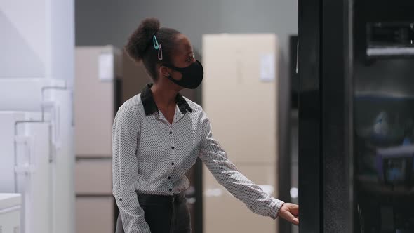Young Black Woman with Face Mask is Choosing Refrigerator with Freezer in Home Appliance Store alt