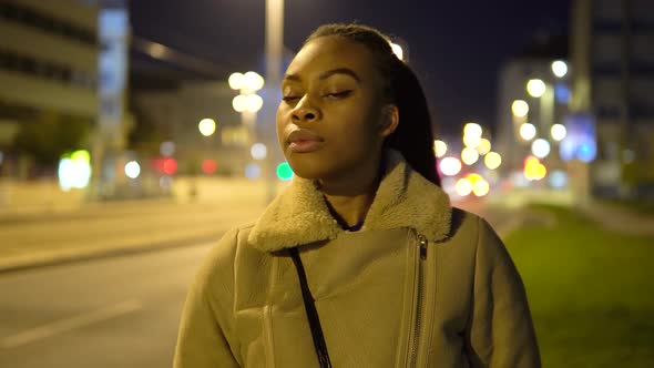 A Young Black Woman Looks Around in a Street in an Urban Area at Night alt