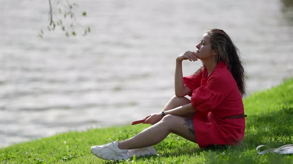 Female City Dweller is Resting on River or Lake Coast in Park in Daytime Enjoying Nature alt