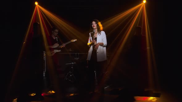 A Stylish Girl Vocalist Sings on Stage in a Vintage Microphone. In the Background Musicians alt