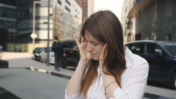 A Woman Sits and Massages the Temples for Headaches on the Background of Skyscrapers alt