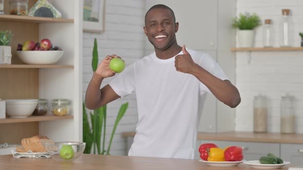 Sporty African Man Showing Thumbs Up While Holding Apple in Kitchen alt