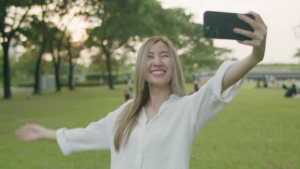 Happy Asian females taking selfie photos on smartphones at a public park during sunset.