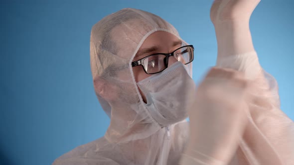 Man in Protective Suit and Medical Mask Puts Rubber Blue Gloves on His Hands