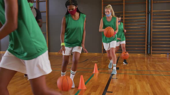 Diverse female basketball team and coach wearing face masks practice dribbling ball alt