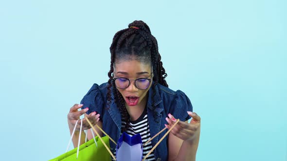 Cute Cheerful Young African American Girl in Blue Clothes Looks in Laminated Cardboard Bags alt