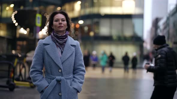 a Woman in a Light Coat and Scarf Walks Along a City Street at Dusk Against a Background of Blurred alt