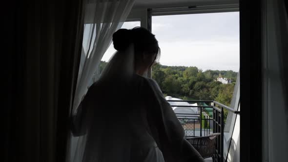 Lovely Bride in White Boudoir Dress Goes To Balcony and Opens Curtains in Hotel Room or Apartment alt