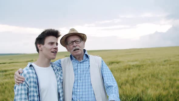 Happy Senior Farmer Stands in Embraces with Son in Barley Field and They Singing alt