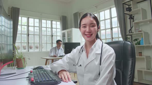 Young Asian Woman Doctor Is Using Computer, She Turns And Smiles To Camera In Workplace alt