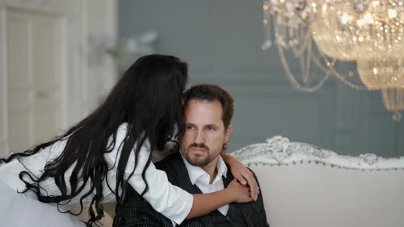 a Bride with Long Dark Hair is Hugging an Unshaven Groom in a White Shirt and Suit on a Light Sofa alt