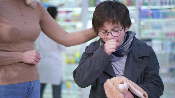 Unwell Caucasian Boy Coughing Standing in Pharmacy with Unrecognizable Woman Caressing Hair alt