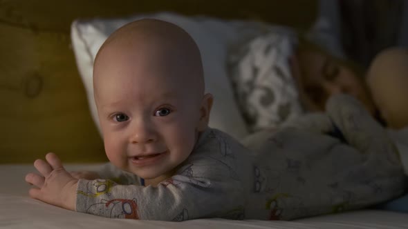 happy baby lies on bed next to sleeping mom in the evening