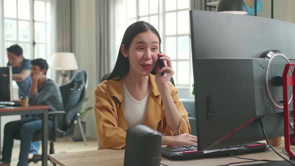 Asian Female Talking On Mobile Phone While Works On Her Personal Computer With Big Display In Office alt
