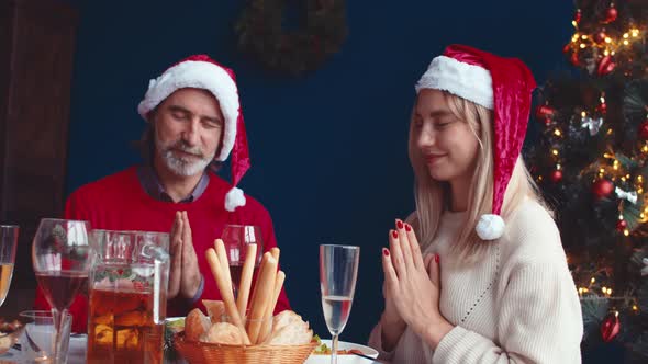 Young Woman and an Adult Man in Christmas Hats Sit at Table with Their Hands Folded and Pray Before alt