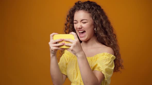 Young Curly Woman Playing Games on Smartphone Against Yellow Background