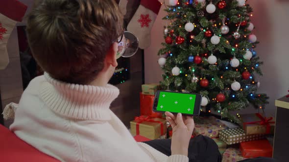 A man enjoying wine and scrolling Green Screen Mockup phone in a Chrismas decorated living room. alt