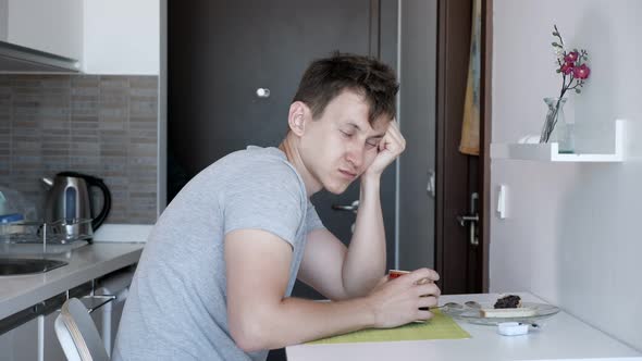 Sleepy Man Is Having a Cup of Tea Sitting at Small Kitchen in Renting Apartment on Vacation. alt
