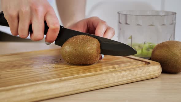 Female Hands Cut the Kiwi with a Knife on 2 Half on a Wooden Board alt