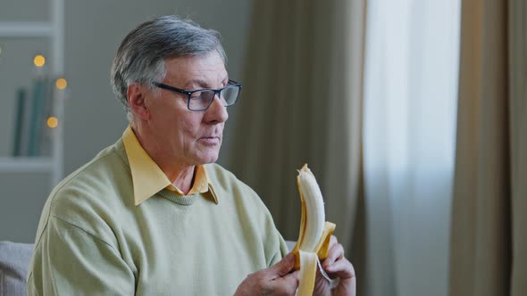 Caucasian Old 60s Male Pensioner Grandpa in Glasses at Home Sitting on Couch Eating Tasty Fresh alt