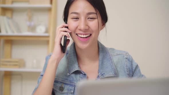 Asian woman working on laptop and talking on phone  while sitting on table in creative office. alt