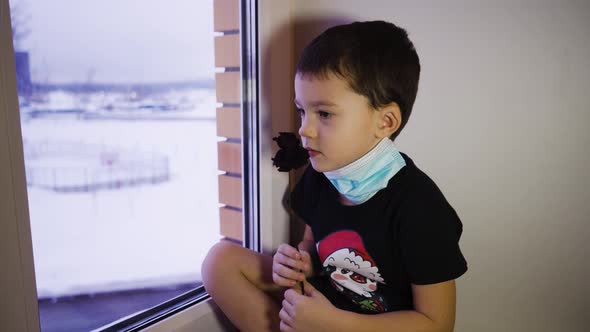 A Sad Child in a Mask at the Window Sniffs a Dried Flower