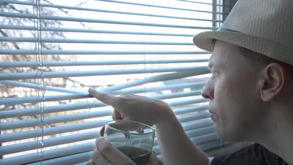 A Young Man in a Hat with Interest Examines What is Happening on the Street Drinks From a Glass alt
