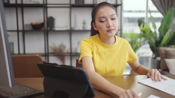 Asian woman sitting at home working at her desk with documents and computer alt